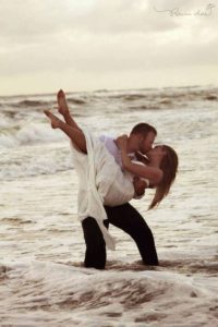 Couple in wedding clothes as he holds her in the beach waves.